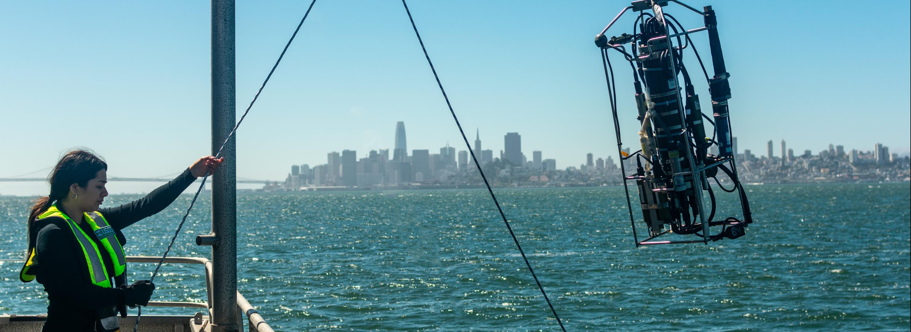 Scientist in yellow vest holds rope while deploying a scientific instrument off a boat with San Francisco skyline in the background