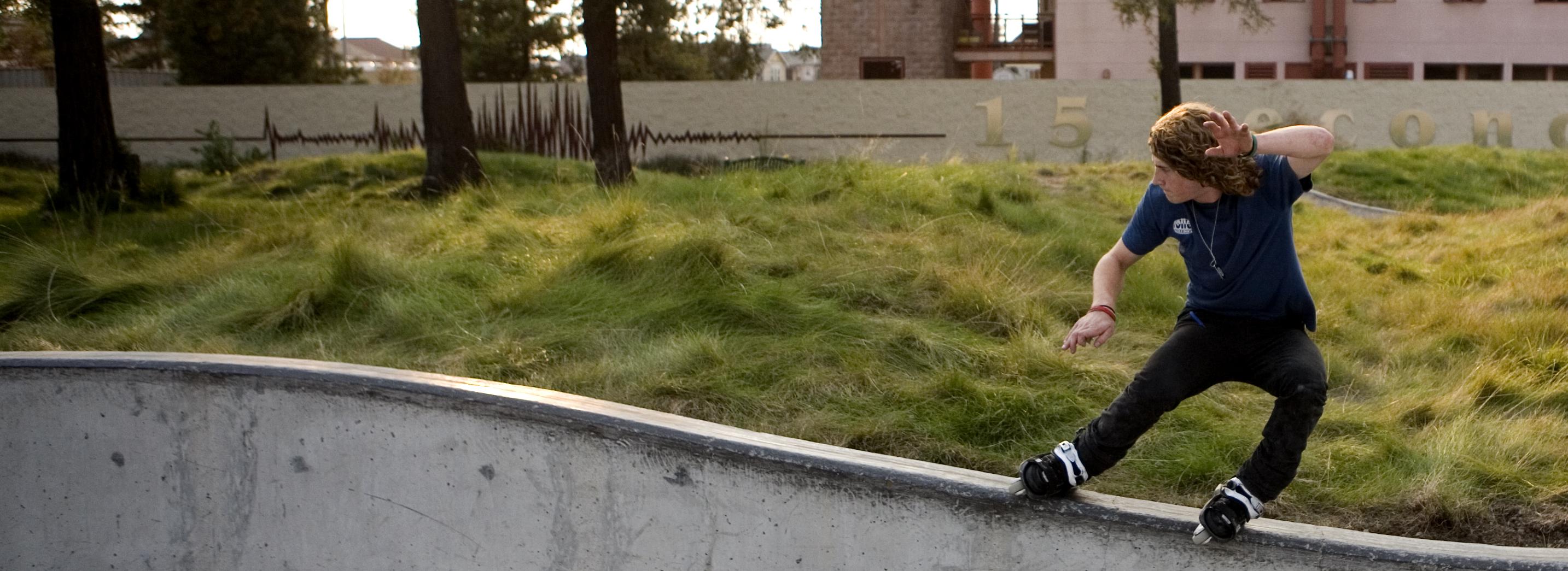 A roller blader rides across a short concrete retaining wall with native landscaping behind them.