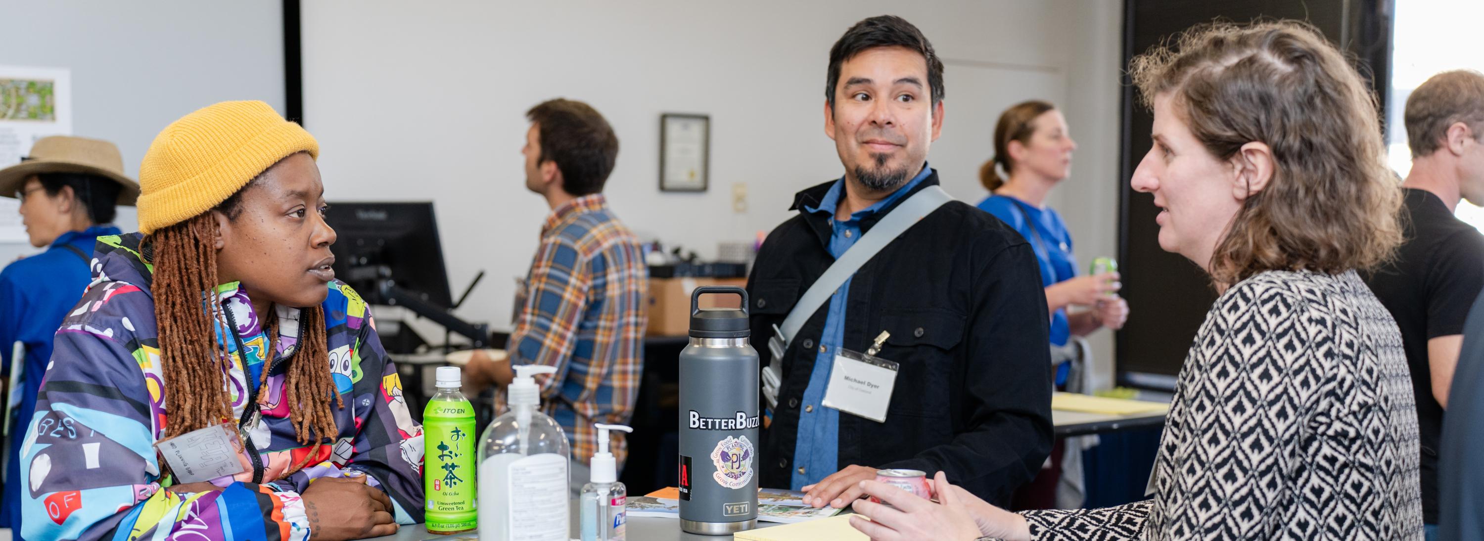  3 people stand chatting around a table with water bottles and post it notes.