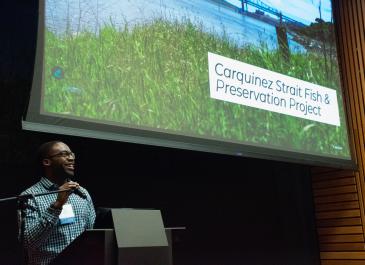 William Ware holds microphone standing behind podium and smiles with Carquinez Strait Fish and Preservation Project slide displayed behind him.