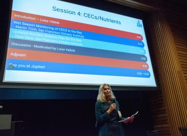 Luisa Valiela stands in front of a large agenda slide titled "Session 4: CECs/Nutrients" while holding a microphone in one hand and a sheaf of notes in the other.
