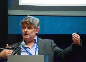 Close-up of David Senn behind a podium gesturing widely and looking towards the side.