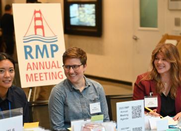 From left to right, Michelle Azurin, Kara Mirmelstein, and Anna de Lopez sit behind a check-in table with a large posterboard behind them with logo and text reading "RMP Annual Meeting." 