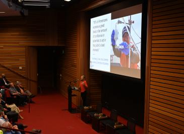 Sierra Garcia presents standing behind podium and gestures with a slide reading "76% of Americans express a great deal or fair amount of confidence in scientists to act in the public’s best interests."