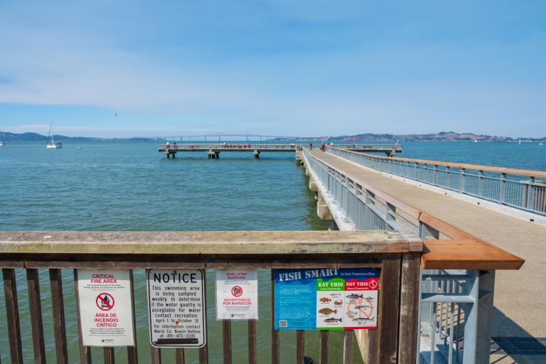 Photo showing a fishing pier in Tiburon with the Bay in the background.  At the entrance to the pier is a sign based on the fish consumption advisory for the Bay.