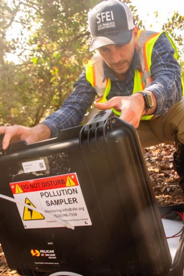 Scientist wearing yellow hazard vest and SFEI baseball cap inspects a black case labeled "Pollution Sampler" under green foliage in the background