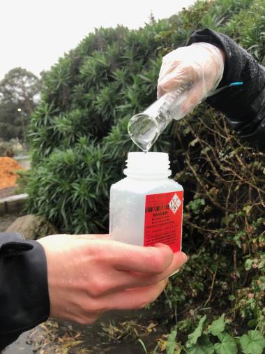 A gloved hand holds a plastic sampling bottle with a red label while another person's gloved hands pours water into it from a graduated cylinder. They are outdoors and in rain gear