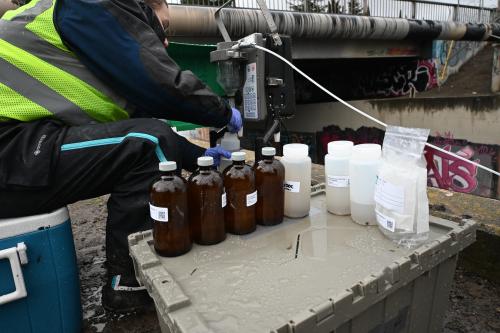 Scientist in green hazard vest and jacket sits on a cooler while filling a bottle with a stormwater sample beside a bridge. Several other dark brown glass and opaque plastic bottles with sampling labels are on top of another cooler in foreground