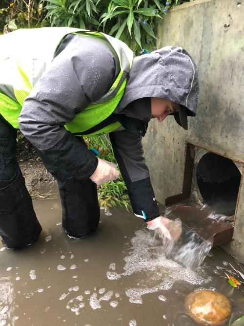 Person in bright green hazard vest and grey hoodie bends down with a bottle to collect stormwater flowing out of a hole in concrete wall at Visitacion Valley
