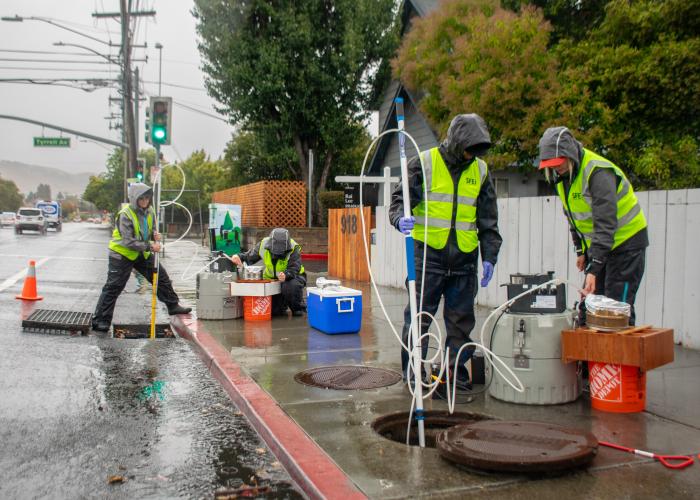 Four people in hazard vests and rain gear use sampling equipment including long poles at an uncovered curbside drainage system while stormwater flows down the road towards the drain