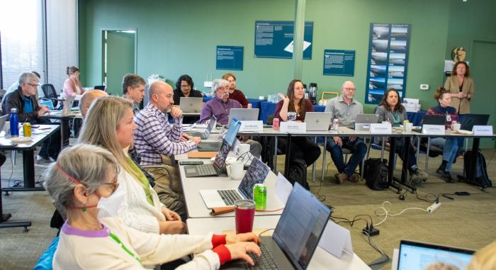 L-joined tables inside a room with people with name placards, laptops, and notes; others are seated in the background