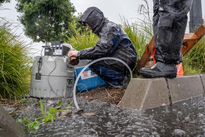 Scientist in rain coat crouches next to water sampling pump besides large tufts of grass. A tube leads to rainwater pooling in the foreground.