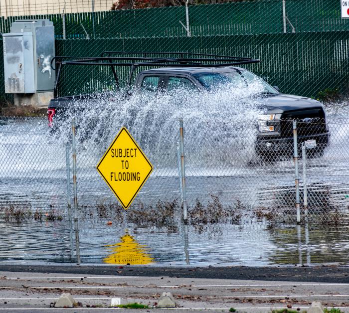 Truck splashes water driving down flooded road behind sign that reads "subject to flooding" on a fence