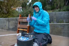 Ezra Miller in blue raincoat kneels on sidewalk and smiles while pointing to graduated cylinder full of water
