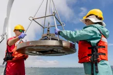 Two scientists in hard hats and life vests hold the edges of a large metal instrument at the edge of a boat