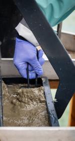 Gloved hand scooping some mud out of a large metal claw-grab style instrument filled with mud