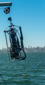 Scientist in yellow vest holds rope while deploying a scientific instrument off a boat with San Francisco skyline in the background