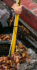 Hand grips a long pole inserted into an open curbside street drain with water flowing into it and