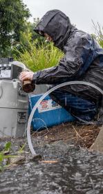 Scientist in rain coat crouches next to water sampling pump besides large tufts of grass. A tube leads to rainwater pooling in the foreground.