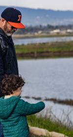 Adult in SF Giants baseball cap holds hand of a child pointing towards the Bay water at Shoreline Park. Photo courtesy of the City of Mountain View. 