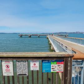 Photo showing a fishing pier in Tiburon with the Bay in the background.  At the entrance to the pier is a sign based on the fish consumption advisory for the Bay. 