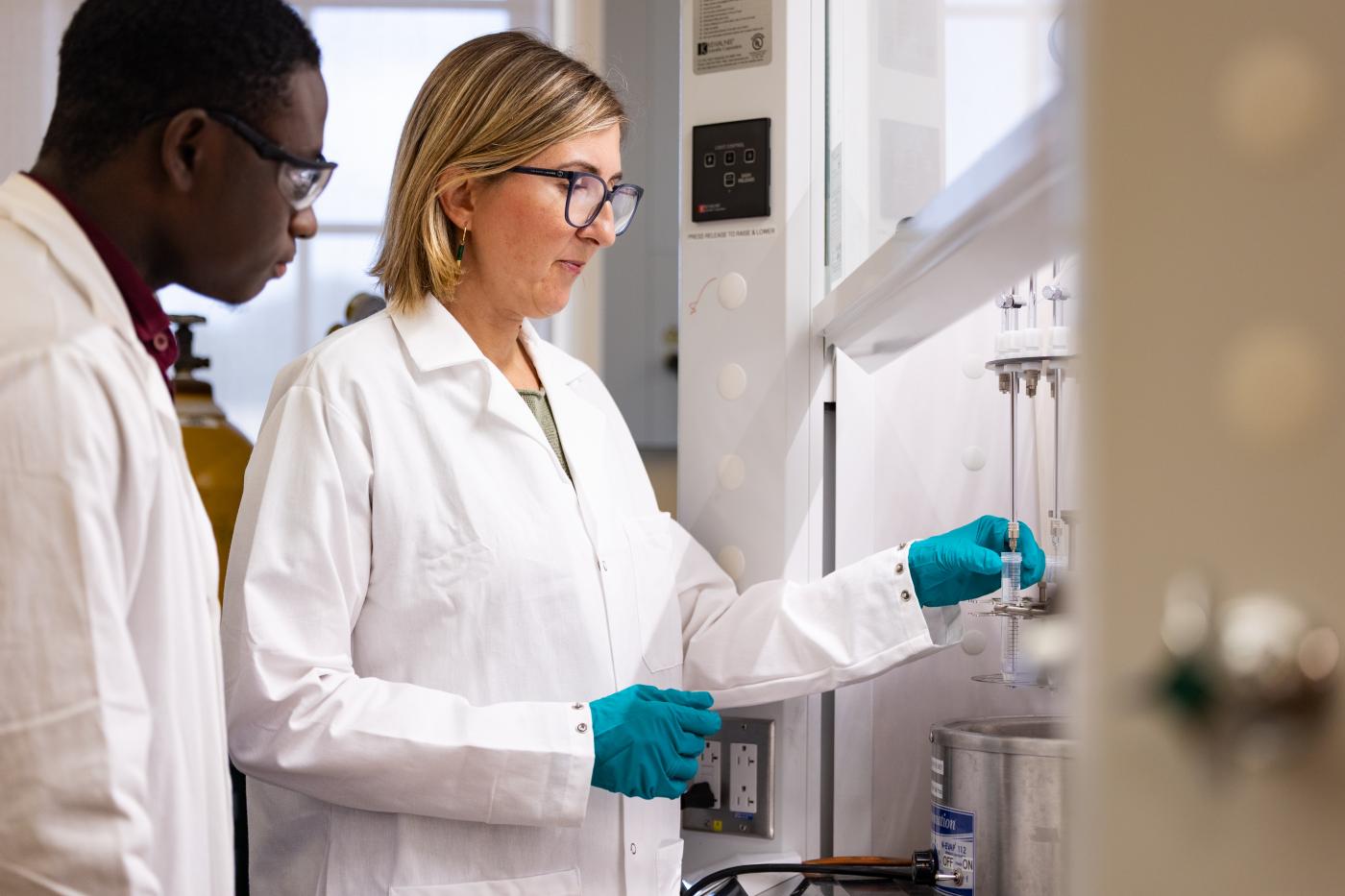 A scientist in lab coat, protective glasses, and gloves hold a gradduated cylinder with clear liquid and a probe inserted while another scientist watches