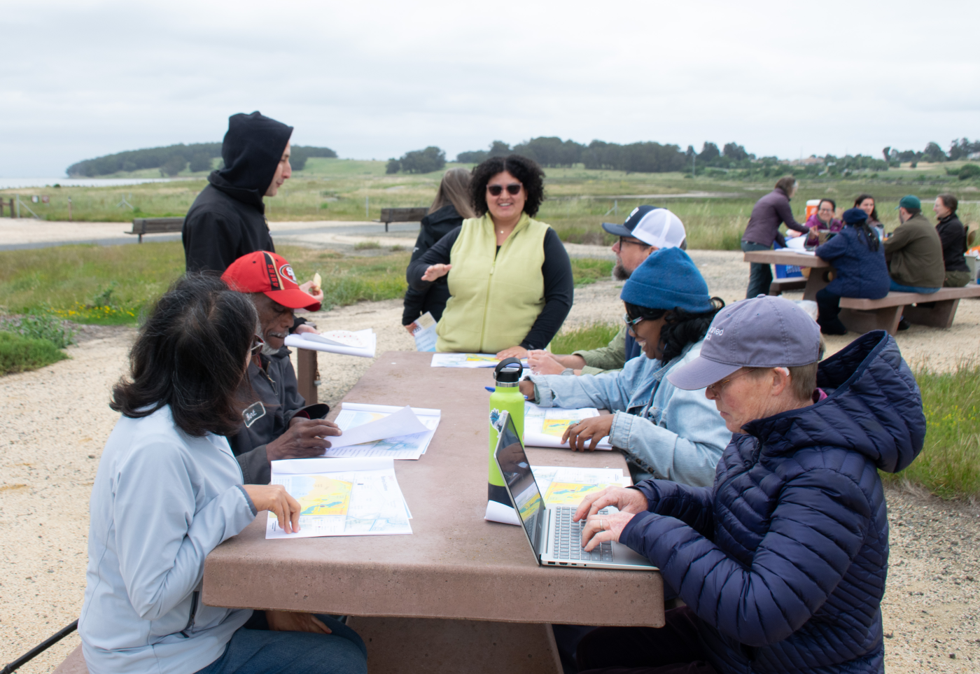 People sit at a park picnic table looking at maps, with woman standing talking to group. In the background the park is surrounded by wetlands.