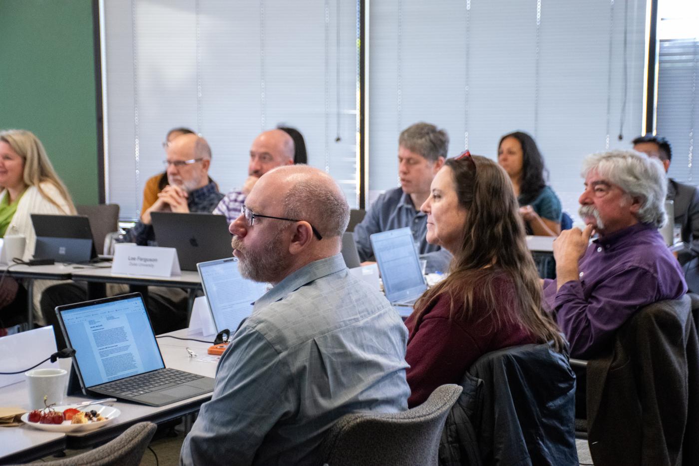 A group of people with laptops sit at a table listening to a speaker.